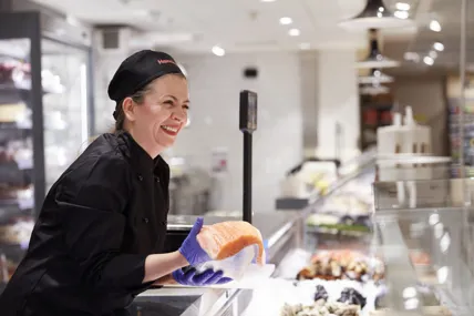 En glad kvinnlig medarbetare i svarta kläder på Hemköp Deli sträcker fram en bit färsk fisk över disken