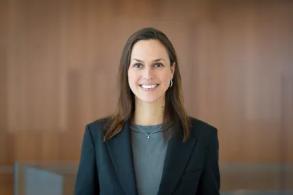 Kvinna i svart kavaj mot en brun trävägg tittar in i kameran